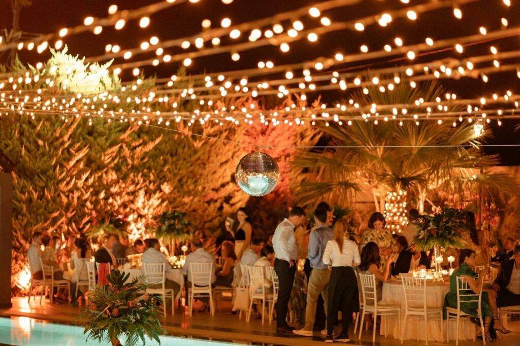 A picture of a Greek wedding showing a disco ball hung above a pool, with fairy lights hung above and lit up trees in the background